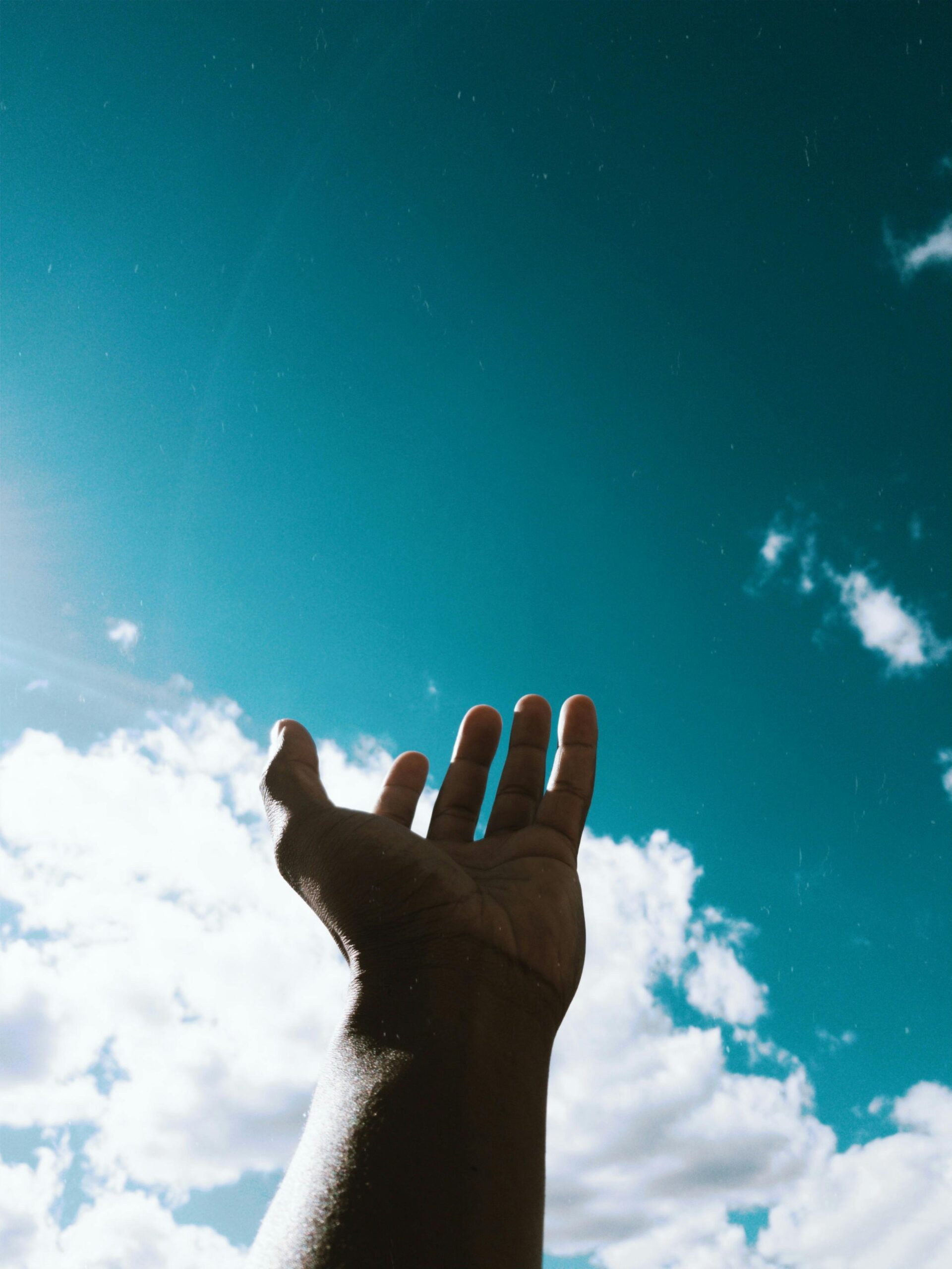 A hand reaching out to a bright sky with scattered clouds, symbolizing hope and aspiration.